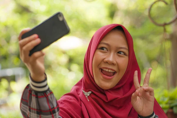 Estilo de vida estudantil muçulmano - jovem bela menina estudante asiática feliz e alegre Indonésia vestindo Islã tradicional hijab cabeça cachecol tomando selfie com telefone celular — Fotografia de Stock