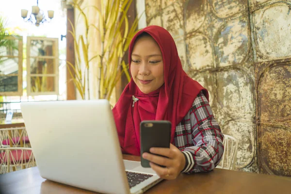 Мусульманская студентка в хиджабе работает с ноутбуком. Молодая счастливая и красивая азиатская индонезийская женщина в исламе головной шарф работает в кафе с помощью компьютера и мобильного телефона — стоковое фото