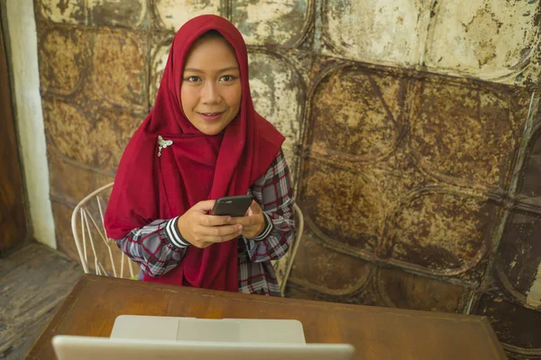 Moslim student meisje in hijab netwerken met laptop. Jonge gelukkige en mooie Aziatische Indonesische vrouw in de islam hoofd sjaal werken in café met behulp van computer en mobiele telefoon — Stockfoto