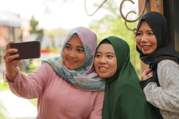 Мусульманка и ее дочери-подростки. Счастливые азиатские индонезийские сёстры делают селфи со своей мамой в традиционном исламском хиджабе в праздничные дни — стоковое фото