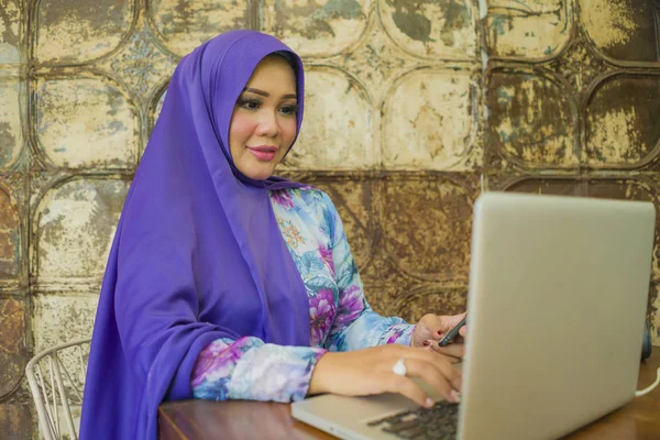 Jovem bela e bem sucedida mulher asiática no tradicional lenço de cabeça hijab trabalhando no computador portátil e telefone celular executando negócios on-line internet relaxado e feliz — Fotografia de Stock