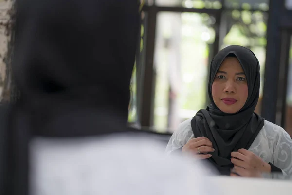 Estilo de vida retrato de mujer musulmana indonesia asiática de mediana edad elegante y feliz en hiyab tradicional ajustando la bufanda islámica frente al espejo — Foto de Stock