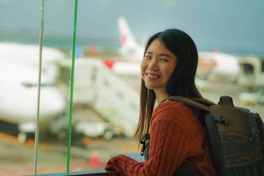 havaalanı kalkış salonunda sırt çantası ile genç mutlu ve heyecanlı Asyalı Japon öğrenci kadın biniş için hazır gülümseyen cam pencereden uçak izliyor