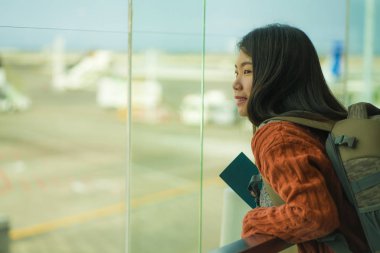 havaalanı kalkış salonunda sırt çantası ile genç mutlu ve heyecanlı Asyalı Çinli öğrenci kadın biniş için hazır gülümseyerek cam pencereden uçak izliyor