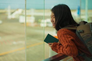 havaalanı kalkış salonunda sırt çantası ile genç mutlu ve heyecanlı Asyalı Koreli öğrenci kadın biniş için hazır gülümseyerek cam pencereden uçak izliyor
