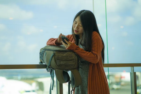 Young beautiful and happy Asian Chinese woman checking mobile phone at airport departure lounge carrying backpack waiting for boarding smiling cheerful ready for holiday travel — Stock Photo, Image