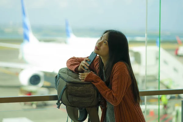 Young beautiful and happy Asian Korean woman checking mobile phone at airport departure lounge carrying backpack waiting for boarding thoughtful and pensive — Stock Photo, Image