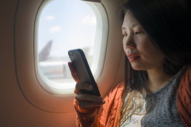 genç mutlu ve güzel Asyalı Çinli turist kadın uçak taşımacılığı ve tatil seyahat el telefonu kullanarak pencerenin yanında uçak oturan heyecanlı gülümseyen