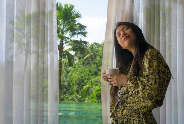 Estilo de vida retrato de la joven hermosa mujer coreana asiática feliz y relajada en elegante albornoz por la ventana de la habitación de hotel de lujo que tiene café por la mañana disfrutando de la vista tropical — Foto de Stock