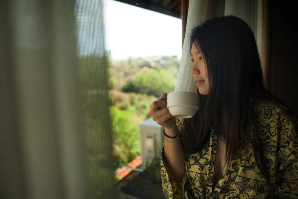 Стиль жизни портрет молодой красивой счастливой и расслабленной азиатской женщины в стильном халате у окна роскошного номера отеля с утренним кофе наслаждаясь тропическим видом — стоковое фото