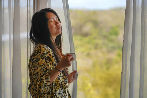 Стиль жизни портрет молодой красивой счастливой и расслабленной азиатской женщины в стильном халате у окна роскошного номера отеля с утренним кофе наслаждаясь тропическим видом — стоковое фото