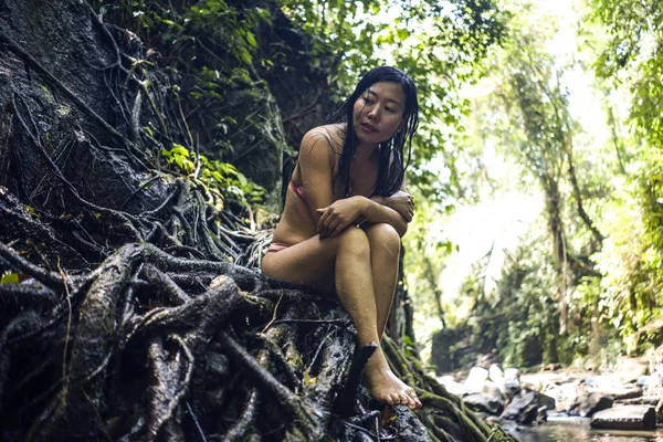 Krásná žena v tropickém začarovaným lese. Mladá atraktivní Čínská dívka v bikinách, která sedí na kmeni v tropické rajské džungli a má tajemný a magický pocit — Stock fotografie