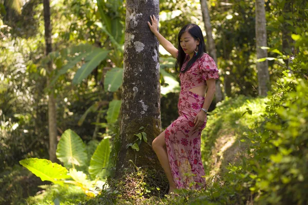 Joven mujer china asiática hermosa y relajada en elegante vestido de verano disfrutando sereno de la belleza de la naturaleza apoyada en el tronco del árbol en la selva tropical fresca — Foto de Stock