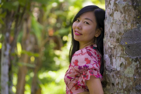 Joven hermosa y relajada mujer china asiática en elegante vestido de verano disfrutando de la serena belleza de la naturaleza apoyada en el tronco del árbol en la selva tropical fresca —  Fotos de Stock