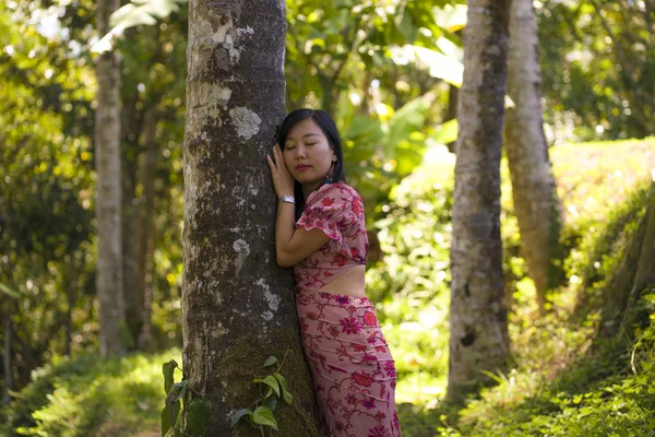 Joven mujer coreana asiática hermosa y relajada en elegante vestido de verano disfrutando serena la belleza de la naturaleza apoyada soñadora en el tronco del árbol en la selva tropical fresca —  Fotos de Stock