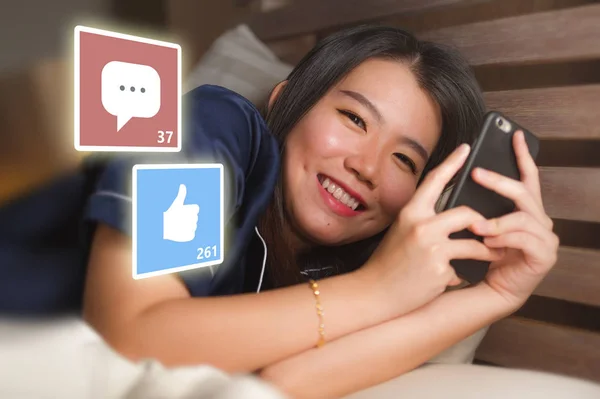 Молодая красивая и счастливая азиатская китаянка в пижаме с помощью мобильного телефона в социальных сетях улыбается весело лежа на кровати дома, получая лайки и комментарии в Интернете — стоковое фото