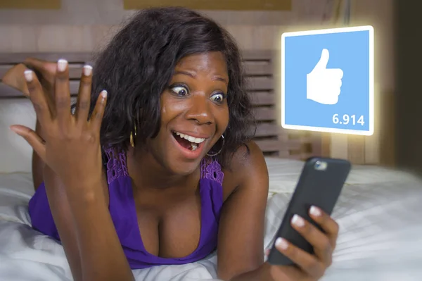 Young beautiful and happy black African American woman in pajamas using mobile phone social media excited and cheerful lying on bed at home receiving lot of likes — Stock Photo, Image
