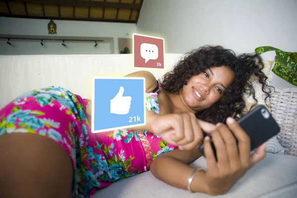 Internet social media app likes y chat comentarios iconos compuesto en joven hermosa y feliz mixta etnia latina afroamericana mujer en casa sofá usando teléfono móvil —  Fotos de Stock