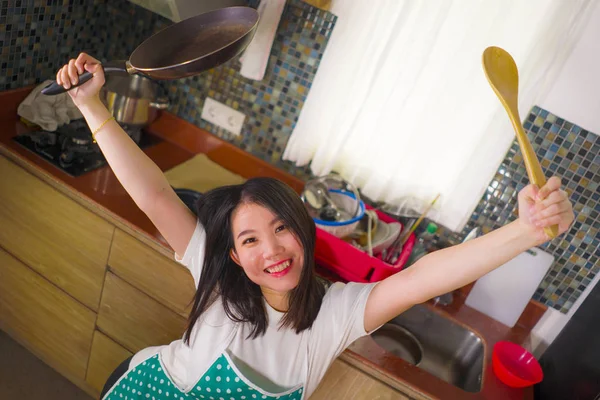 Молодая красивая и счастливая азиатская китаянка в кухонном фартуке держит кастрюлю и ложку улыбаясь весело наслаждаясь домашними делами повар и уборщица позирует игриво — стоковое фото