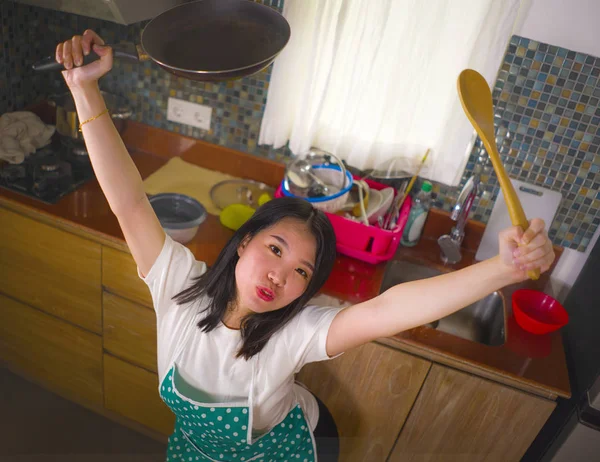 Jovem bonita e feliz mulher asiática coreana na cozinha avental segurando panela e colher sorrindo alegre desfrutando de tarefas domésticas cozinhar e limpeza posando brincalhão — Fotografia de Stock