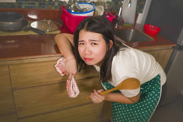 Young tired and stressed Asian Chinese woman in cook apron working leaning on kitchen sink lazy in moody and upset face housekeeping at home and crying — Stock Photo, Image