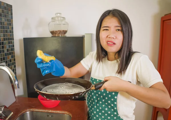 Domestic chores lifestyle portrait of young tired and stressed Asian Chinese woman in cook apron washing dishes at kitchen sink working lazy in moody and upset face housekeeping — Stock Photo, Image