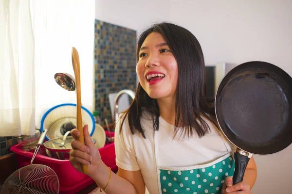 Jovem bela e feliz mulher chinesa asiática em casa em avental segurando panela de cozinha e colher sorrindo brincalhão desfrutando de tarefas domésticas — Fotografia de Stock