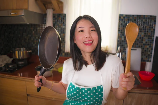Jonge mooie en gelukkige Aziatische Chinese vrouw thuis in schort houden kookpan en lepel speelse glimlachend genieten van huishoudelijke klusjes — Stockfoto