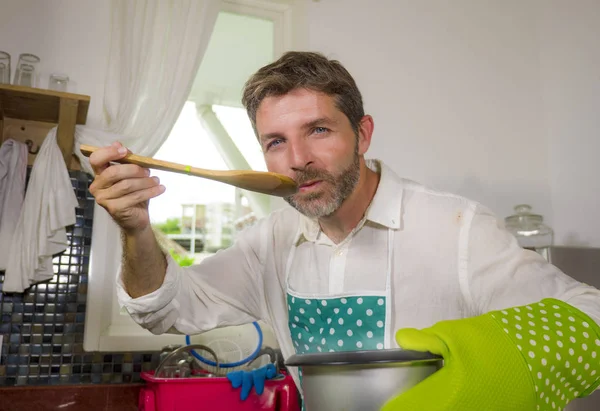 Porträt eines glücklichen und attraktiven Mannes in Schürze und Handschuh, der Kochtopf hält und mit einem Löffel den köstlichen Geschmack der Suppe in der erfolgreichen Hausmannskost verkostet — Stockfoto