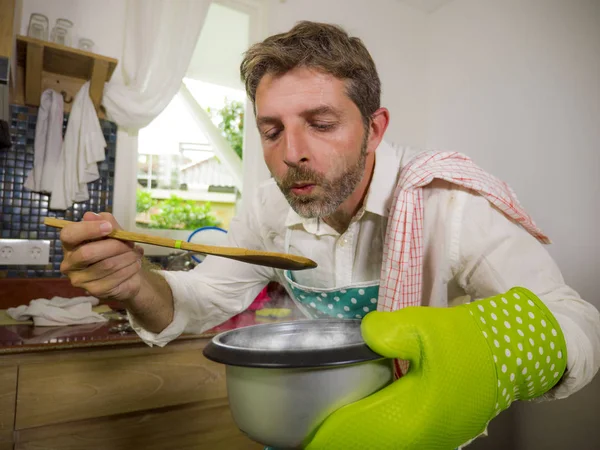 Domestic lifestyle portrait of happy and attractive man in apron and glove holding cooking pot tasting with spoon the delicious flavour of soup in successful home cook — Stock Photo, Image