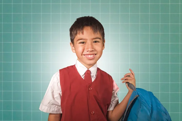 Ung pojke 7 eller 8 år gammal i väst och halsduk enhetlig hållande väska leende glada framför gröna svarta tavlan upphetsad om tillbaka till skolan i barn utbildning Concept — Stockfoto
