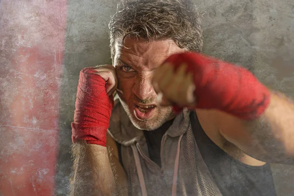 Estilo de vida gimnasio retrato de joven atractivo y feroz hombre entrenamiento boxeo en el club de fitness haciendo entrenamiento de perforación de bolsas pesadas con envolturas de muñeca en aspecto de luchador rudo — Foto de Stock