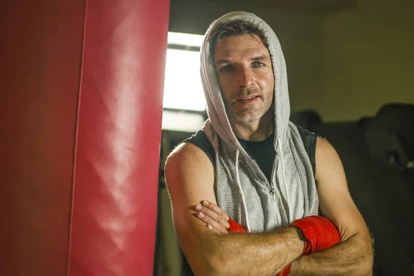 sport fitness lifestyle portrait of young happy and sweaty man boxing at gym working out sweaty in hoodie vest training on heavy bag looking coo
