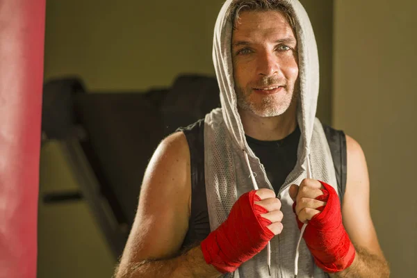 young happy and sweaty man boxing at gym working out sweaty in hoodie vest training boxing on heavy bag looking cool smiling positive in fighter wrist wraps