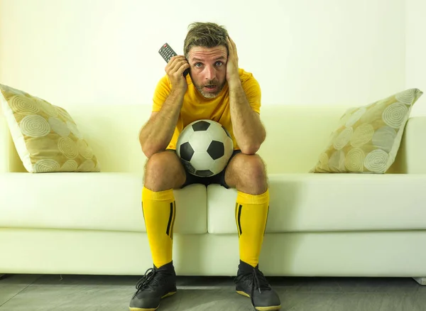 在电视上看足球比赛的年轻而紧张又兴奋的足球迷穿着队服坐在沙发上，穿着队服，感受压力和情绪的滑稽画像 — 图库照片