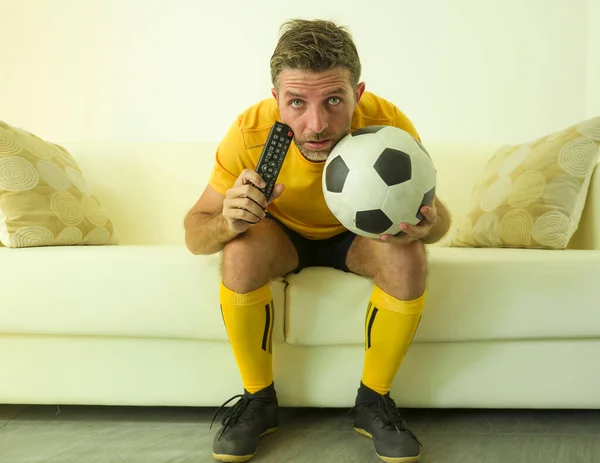 funny portrait of young stressed and excited football fan man watching soccer game on TV at home couch dressed in team player uniform feeling  stress and emotion