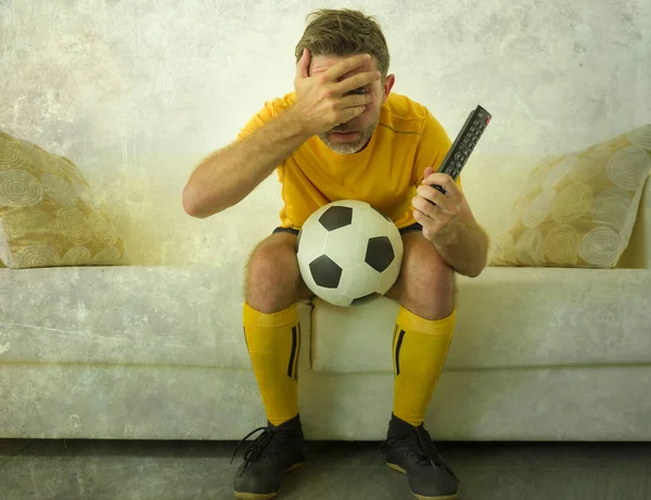 funny portrait of young stressed and excited football fan man watching soccer game on TV at home couch dressed in team player uniform feeling  stress and emotion