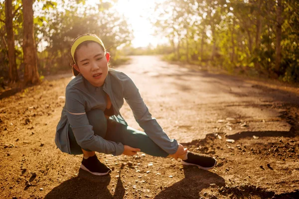 Fitness estilo de vida retrato de jovem atraente mulher corredor asiático sofrendo lesão esportiva durante jogging treino na estrada por do sol segurando tornozelo torcido na dor — Fotografia de Stock