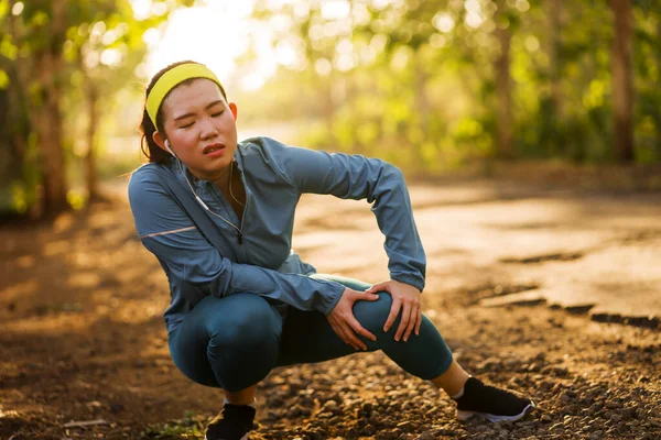 Fitness estilo de vida retrato de jovem atraente mulher corredor asiático sofrendo lesão esportiva durante jogging treino na estrada por do sol segurando joelho na dor — Fotografia de Stock