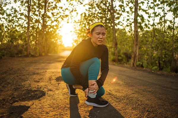 Fitness lifestyle portrait of young attractive Asian runner woman suffering sport injury during jogging workout on sunset road holding twisted ankle in pain — Stock Photo, Image