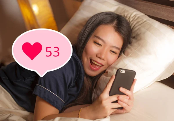 Молодая красивая и счастливая азиатская девушка получает лайки в социальных сетях. Великолепная китаянка в постели по ночам радостно улыбается в интернет-приложении — стоковое фото