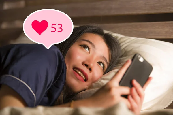 Молодая красивая и счастливая азиатская девушка получает лайки в социальных сетях. Великолепная кореянка в постели ночью взволнована получением положительных отзывов в интернет-приложении улыбаясь весело — стоковое фото
