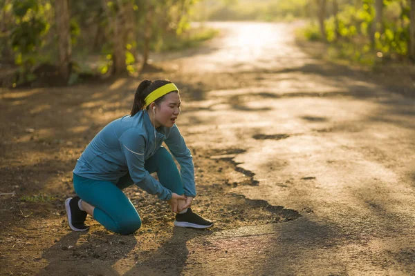 Fitness styl życia portret młodej atrakcyjnej azjatyckiej biegaczki cierpiącej kontuzję sportową podczas treningu joggingowego na zachodzie słońca trzymającej skręconą kostkę w bólu — Zdjęcie stockowe