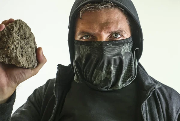 young man as radical and aggressive anarchist rioter holding brick . furious anti-system protester in face mask throwing stone in violent riot  isolated on white