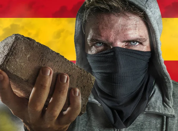 Young man as ultra and extremist Spanish nationalism defender. violent protester in face mask holding a brick at fighting riot in radical demonstration isolated on Spain flag — Stock Photo, Image