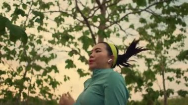 Yavaş çekim, genç ve sağlıklı Asyalı bir kadının günbatımında ağaçların arasında koşup güzel kırsal yolda koşu yaparken görüntüsünü takip ediyor. 