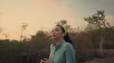 Genç ve sağlıklı Asyalı bir kadının günbatımında ağaçların arasında koşup güzel kırsal yolda koşu yaparken görüntülerini izliyoruz. 