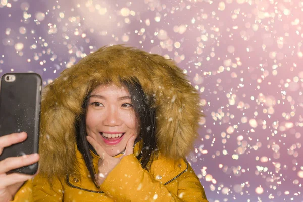 Joven hermosa y feliz mujer china asiática en la capucha chaqueta de invierno tomando retrato selfie con el teléfono móvil al aire libre bajo la nieve sonriente alegre disfrutando de la Navidad — Foto de Stock