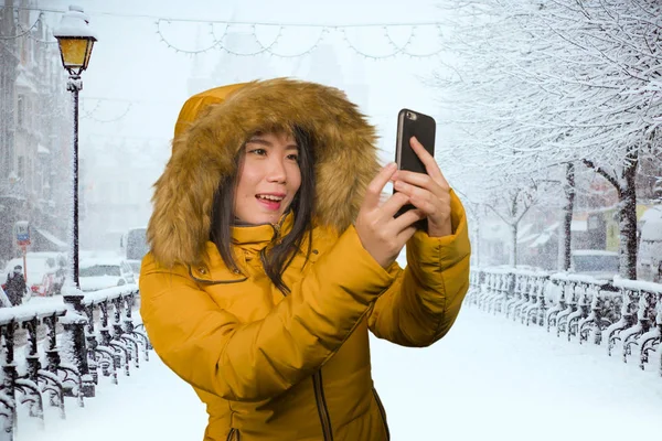 Joven hermosa y feliz mujer asiática coreana en la capucha chaqueta de invierno tomando retrato selfie con el teléfono móvil al aire libre bajo la nieve sonriente alegre disfrutando de la Navidad — Foto de Stock