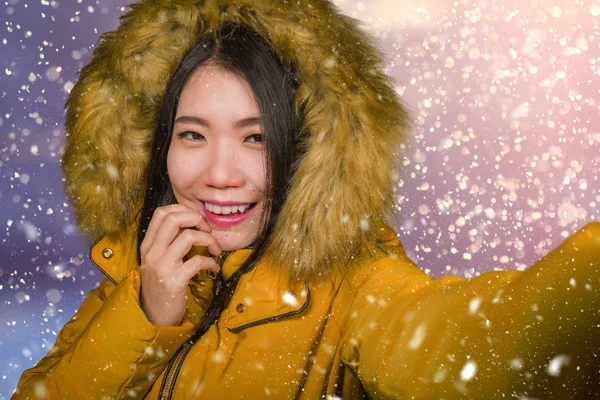 Jovem bonita e feliz mulher coreana asiática no capuz casaco de inverno tendo retrato selfie com telefone celular ao ar livre sob a neve sorrindo alegre desfrutando de Natal — Fotografia de Stock